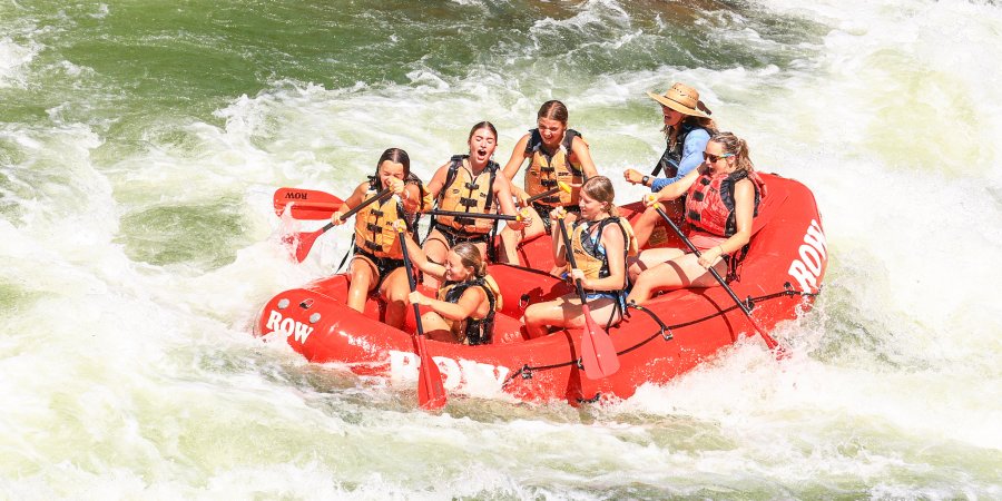 A red ROW branded raft in the middle of a rapid full of guests smiling and paddling