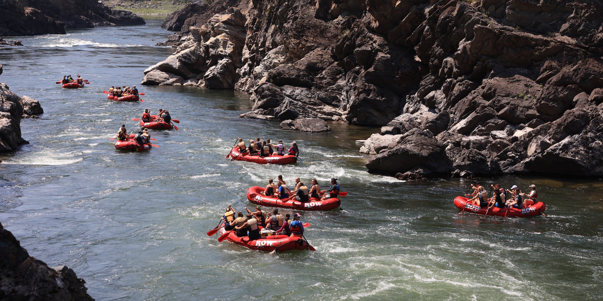 Multiple rafts floating down a scenic river canyon with rocky cliffs.