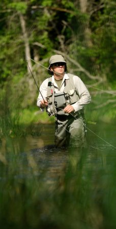Fly fisherman wading in the St. Joe River in Idaho, casting a line through clear, shallow water surrounded by lush forest.