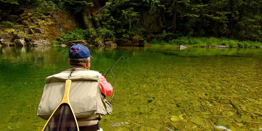 Fly fisherman casting into a crystal-clear Idaho river surrounded by rocky cliffs and dense forest. I