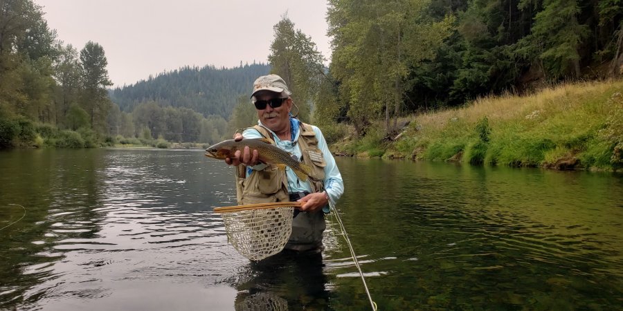  Smiling fly fisherman in waders holding a large cutthroat trout on a clear Idaho stream.