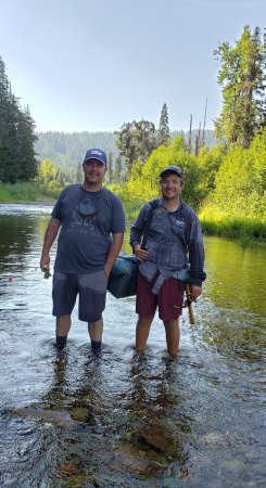Two anglers standing in a shallow Idaho river with fly fishing gear, surrounded by green forest and mountain scenery.